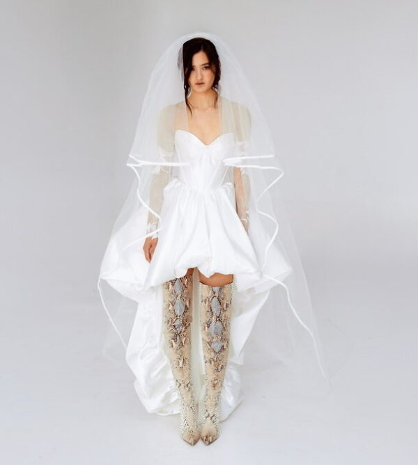Modern high-low wedding dress with corset bodice, detachable lace sleeves, and thigh-high snakeskin boots, perfect for alternative and fashion-forward brides