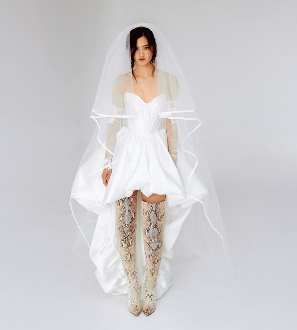 Modern high-low wedding dress with corset bodice, detachable lace sleeves, and thigh-high snakeskin boots, perfect for alternative and fashion-forward brides